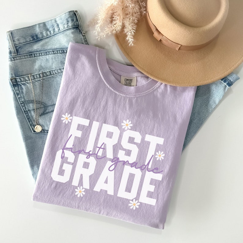 Op de afbeelding: Een lavendelkleurig T-shirt met de tekst "FIRST first grade" in wit met een paarse omtrek. De tekst wordt omringd door kleine witte madeliefjes met gele centra.