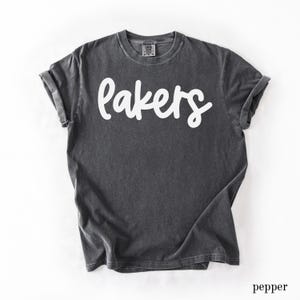 May include: A dark gray t-shirt with the word "Lakers" in white script across the front. The shirt has short sleeves and a crew neck. The bottom right corner of the image has the word "pepper".