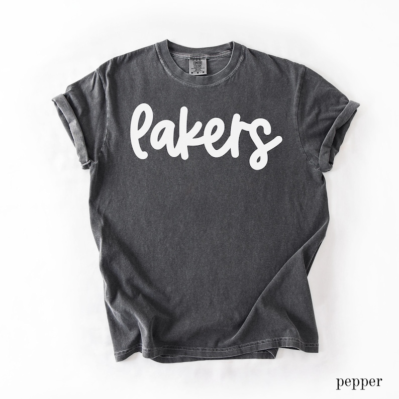 May include: A dark gray t-shirt with the word "Lakers" in white script across the front. The shirt has short sleeves and a crew neck. The bottom right corner of the image has the word "pepper".