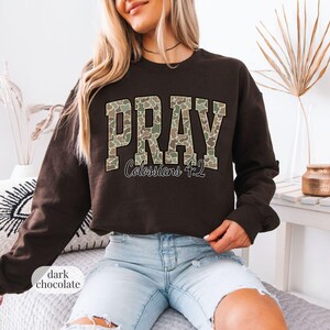 May include: Dark chocolate-colored sweatshirt with the word "PRAY" in a camouflage pattern across the chest. Below the word "PRAY" is the text "Colossians 4:2". The sweatshirt is a long-sleeved crew neck.