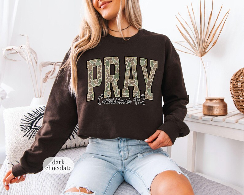 May include: Dark chocolate-colored sweatshirt with the word "PRAY" in a camouflage pattern across the chest. Below the word "PRAY" is the text "Colossians 4:2". The sweatshirt is a long-sleeved crew neck.