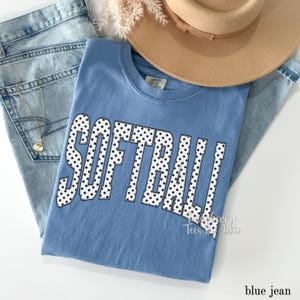 May include: A light blue t-shirt with the word "SOFTBALL" in large white letters filled with black polka dots. The shirt is folded on top of a pair of blue jeans, with a tan hat and a gold necklace visible.