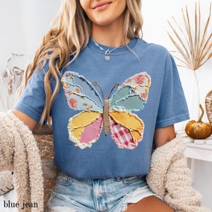 May include: A blue t-shirt featuring a patchwork butterfly design. The butterfly wings are composed of various fabrics in pastel colors, including floral, gingham, and solid patterns. The shirt has short sleeves and a relaxed fit. The text "blue jean" is at the bottom.