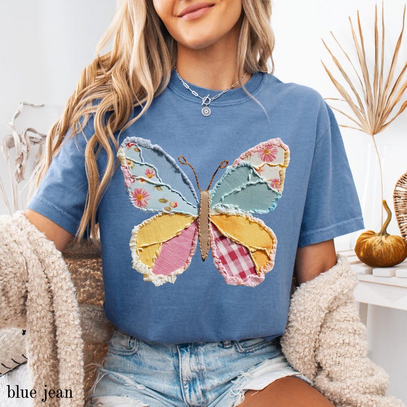 May include: A blue t-shirt featuring a patchwork butterfly design. The butterfly wings are composed of various fabrics in pastel colors, including floral, gingham, and solid patterns. The shirt has short sleeves and a relaxed fit. The text "blue jean" is at the bottom.