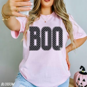 Puede incluir: Una camiseta de manga corta de color rosa claro con la palabra BOO impresa en letras may&uacute;sculas negras grandes. Cada letra est&aacute; decorada con un patr&oacute;n de lunares blancos. La camiseta tiene cuello redondo y un corte holgado y sencillo.