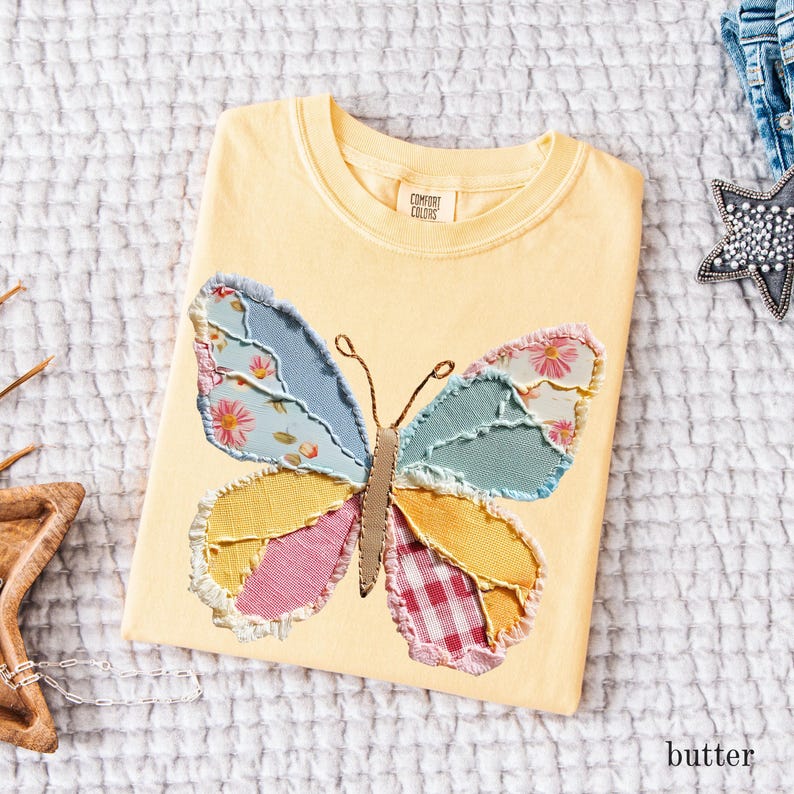 May include: A yellow Comfort Colors t-shirt with a large butterfly applique. The butterfly wings are made of patchwork fabric in shades of blue, pink, yellow, and red. The shirt is folded and lying on a white textured surface. The word "butter" is visible.