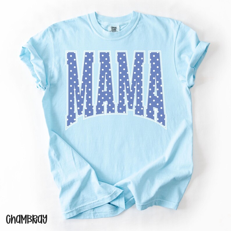 May include: Light blue t-shirt with the word "MAMA" in large, blue, polka-dotted letters. The shirt has short sleeves and a crew neck. The word "Chambray" is printed in the bottom left corner.