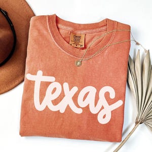 May include: A coral-colored t-shirt with the word "Texas" in a white, cursive font. A gold necklace with a pendant rests on the shirt. A brown hat and dried palm leaves are also in the image.