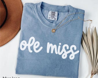 May include: A blue jean colored t-shirt with the words "ole miss" in white script. A gold necklace with a pendant is draped across the shirt. The shirt is folded and has a Comfort Colors tag.