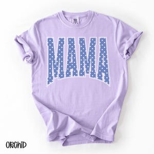 May include: A light purple t-shirt with the word "MAMA" in large, blue letters filled with white polka dots. The shirt has a classic crew neck and short sleeves. The word "ORCHID" is printed in black at the bottom left.