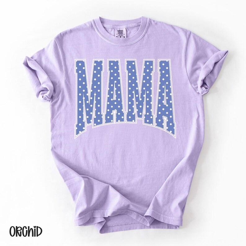 May include: A light purple t-shirt with the word "MAMA" in large, blue letters filled with white polka dots. The shirt has a classic crew neck and short sleeves. The word "ORCHID" is printed in black at the bottom left.