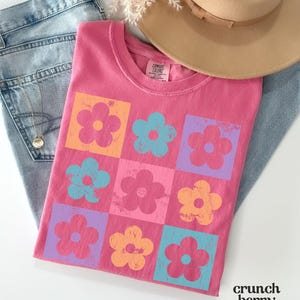 May include: Pink t-shirt with a graphic of a grid of colorful flowers. The flowers are in shades of pink, orange, blue, and purple.