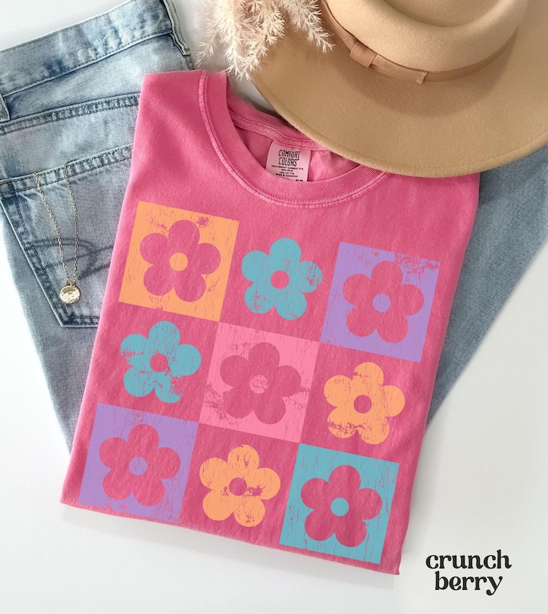 May include: Pink t-shirt with a graphic of a grid of colorful flowers. The flowers are in shades of pink, orange, blue, and purple.