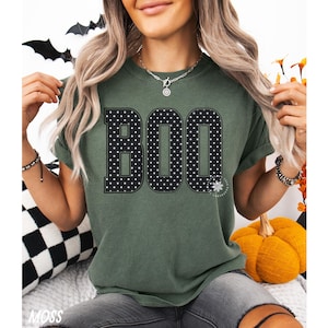 Puede incluir: Una camiseta de manga corta color verde musgo con la palabra BOO impresa en grandes letras negras con un patr&oacute;n de lunares blancos. La prenda tiene cuello redondo y un corte holgado, destacando un dise&ntilde;o gr&aacute;fico centrado en el pecho.
