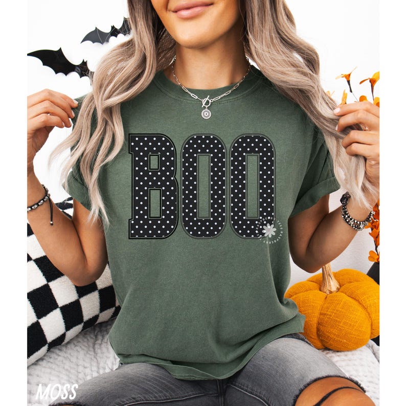 Puede incluir: Una camiseta de manga corta color verde musgo con la palabra BOO impresa en grandes letras negras con un patr&oacute;n de lunares blancos. La prenda tiene cuello redondo y un corte holgado, destacando un dise&ntilde;o gr&aacute;fico centrado en el pecho.