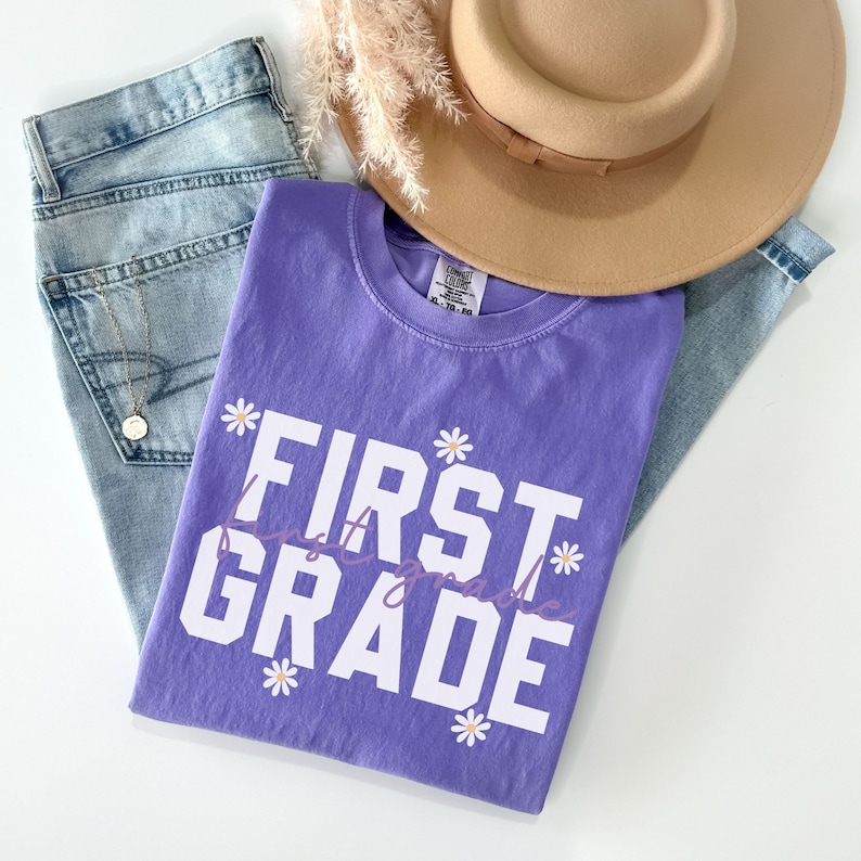 Op de afbeelding: Paars T-shirt met witte tekst die "FIRST GRADE" zegt met kleine paarse bloemen rond de tekst.