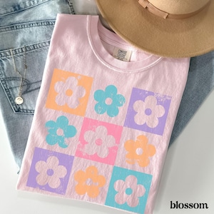 May include: A pink t-shirt with a graphic of a grid of colorful flowers. The flowers are in shades of orange, blue, pink, and purple. The text "COMFORT COLORS" is visible on the shirt.
