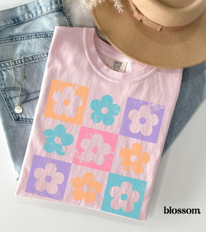 May include: A pink t-shirt with a graphic of a grid of colorful flowers. The flowers are in shades of orange, blue, pink, and purple. The text "COMFORT COLORS" is visible on the shirt.