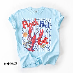 Op de afbeelding: Lichtblauw T-shirt met een afbeelding van een rode kreeft en de woorden "Pinch Peel Eat" in rood en blauw. Het ontwerp bevat een strik, citroenpartjes en een gestreepte achtergrond. Het shirt is gemaakt van een zacht materiaal.