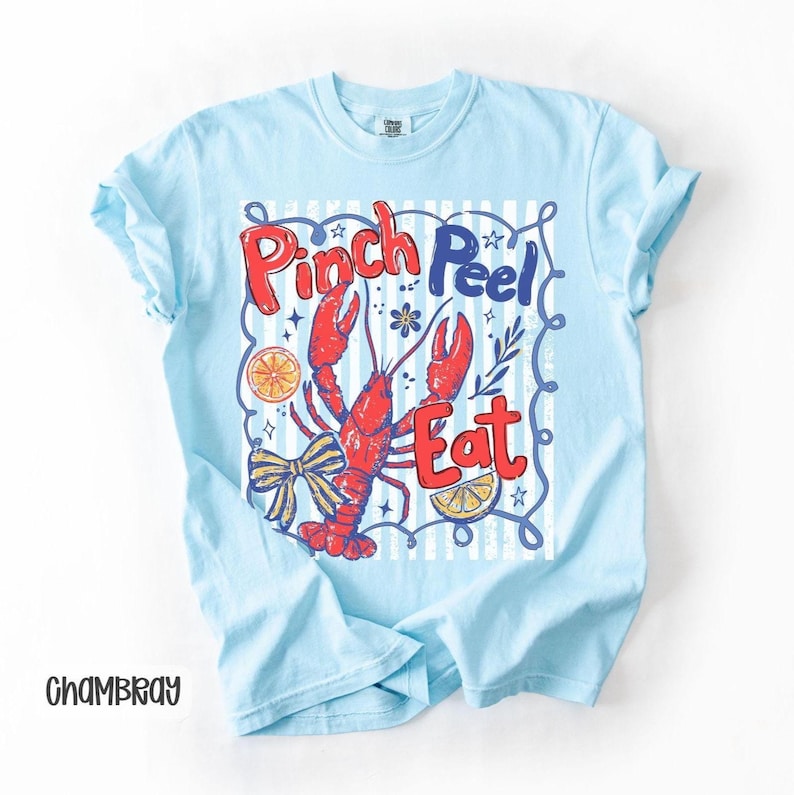 Op de afbeelding: Lichtblauw T-shirt met een afbeelding van een rode kreeft en de woorden "Pinch Peel Eat" in rood en blauw. Het ontwerp bevat een strik, citroenpartjes en een gestreepte achtergrond. Het shirt is gemaakt van een zacht materiaal.