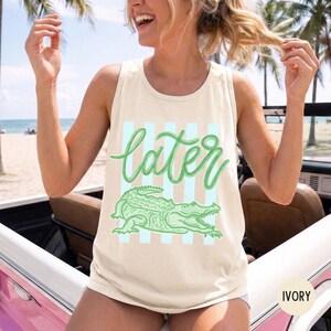 May include: Ivory tank top with the word "later" in green script, over a green alligator illustration and light blue stripes. The tank top is sleeveless and made of a soft material.