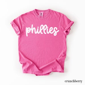May include: A bright pink t-shirt with the word "phillies" in white script across the front. The shirt has rolled-up sleeves and a crew neck. The word "crunchberry" is printed in a small font at the bottom.