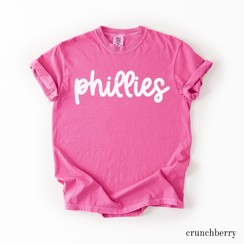 May include: A bright pink t-shirt with the word "phillies" in white script across the front. The shirt has rolled-up sleeves and a crew neck. The word "crunchberry" is printed in a small font at the bottom.