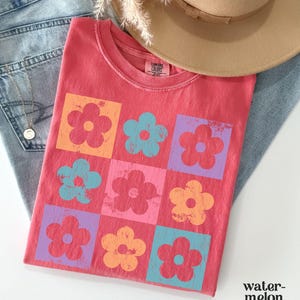 May include: A red t-shirt with a pattern of colorful flowers in squares. The flowers are in shades of pink, blue, orange, and yellow.