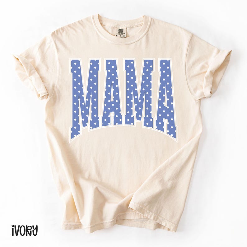 May include: Ivory-colored t-shirt with the word "MAMA" in large, blue, polka-dotted letters. The text has a white outline. The shirt has short sleeves and a crew neck.