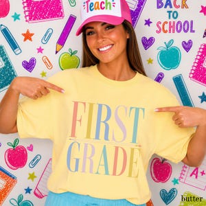 May include: A pale yellow short-sleeve t-shirt featuring the text FIRST GRADE in large, multicolored pastel letters across the chest. The shirt is paired with a pink and white trucker-style hat with the word Teach printed on the front panel.