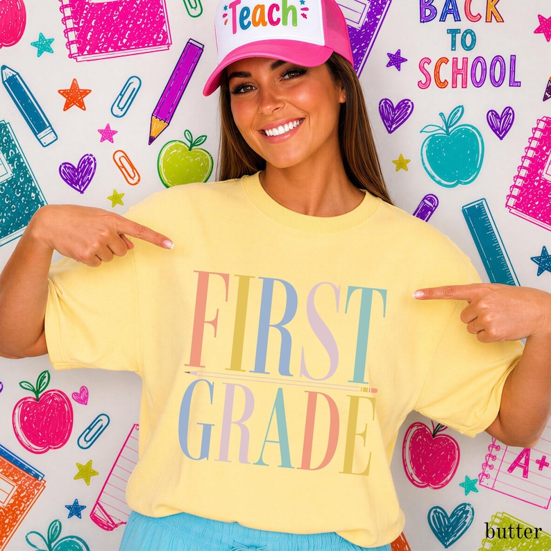 May include: A pale yellow short-sleeve t-shirt featuring the text FIRST GRADE in large, multicolored pastel letters across the chest. The shirt is paired with a pink and white trucker-style hat with the word Teach printed on the front panel.