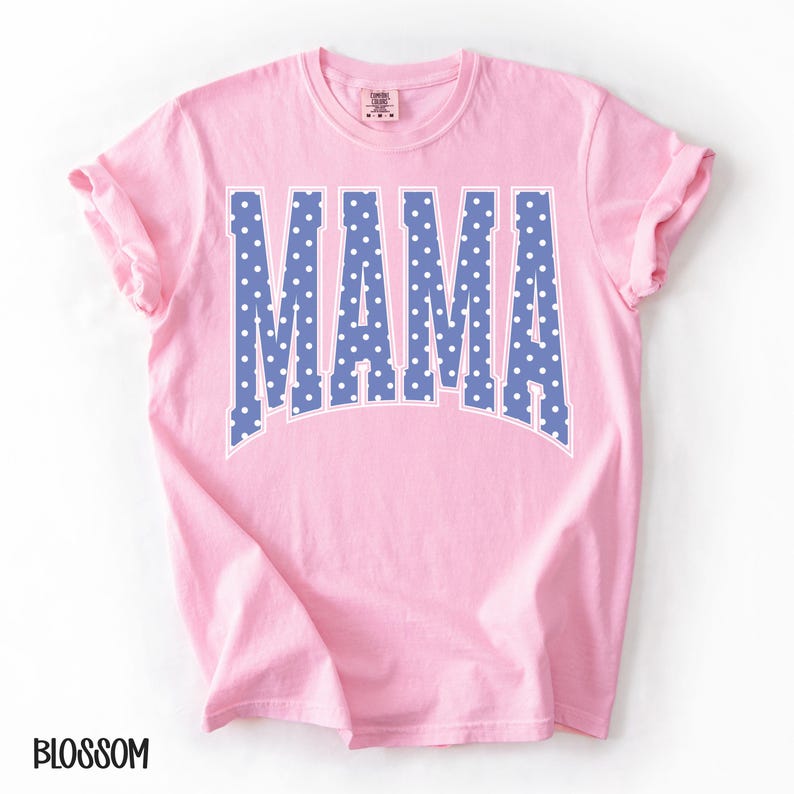 May include: A light pink t-shirt with the word "MAMA" in large, blue, polka-dotted letters. The letters have a white outline. The word "BLOSSOM" is printed in black at the bottom left of the shirt.