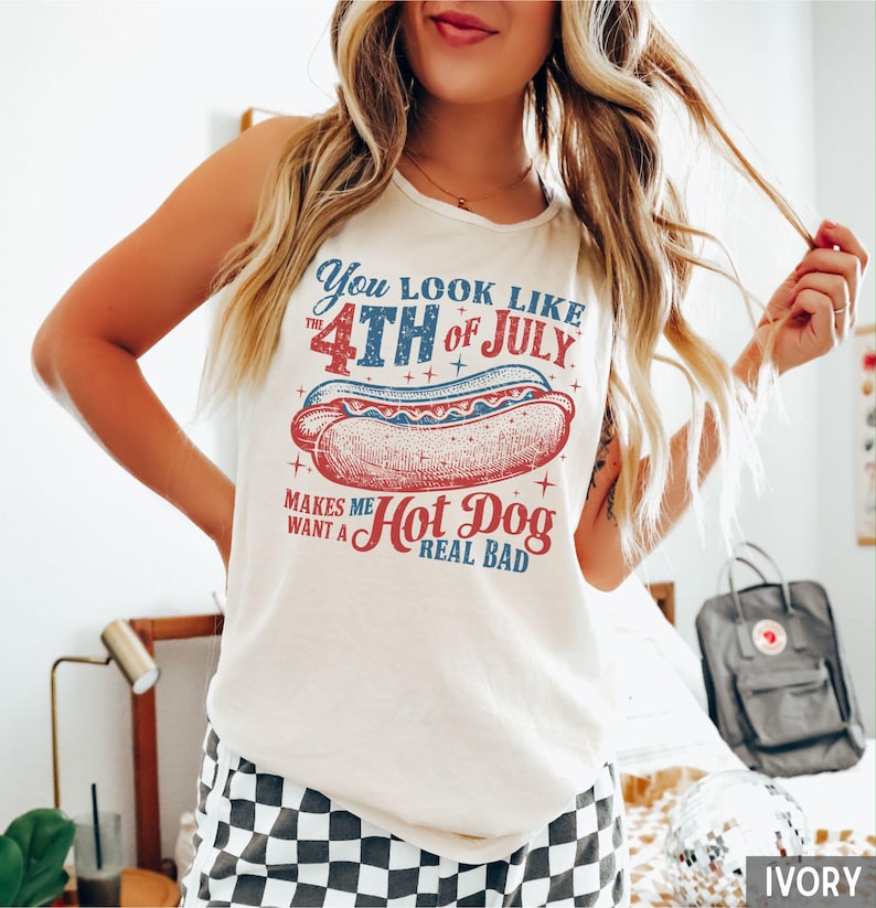 Puede incluir: Camiseta sin mangas color marfil con un gr&aacute;fico que dice "You look like the 4th of July makes me want a hot dog real bad." El gr&aacute;fico presenta una ilustraci&oacute;n de perrito caliente rojo, blanco y azul.