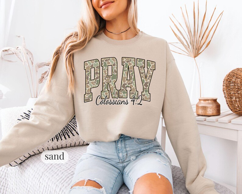 May include: Sand-colored sweatshirt with the word "PRAY" in a camouflage pattern across the chest. The text "Colossians 4:2" is below the word "PRAY". The sweatshirt has long sleeves and a crew neck.