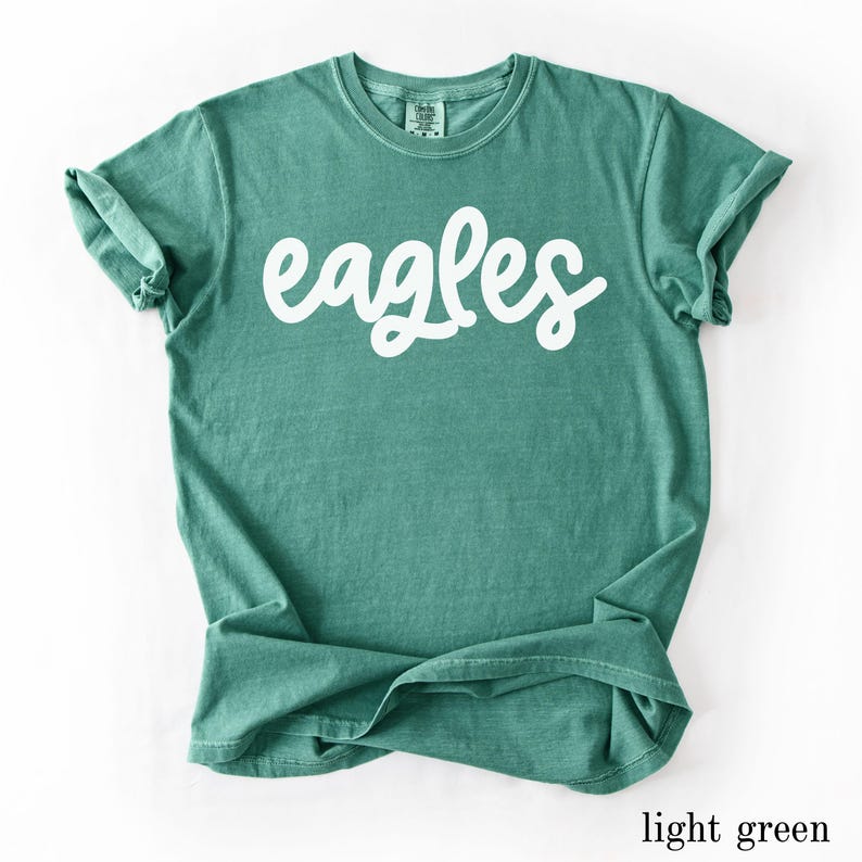 May include: A light green t-shirt with the word "eagles" in white script across the front. The shirt has short sleeves and a crew neck. The bottom of the shirt is slightly folded. The shirt is made of a soft material.