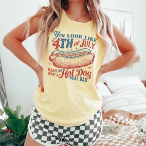 Puede incluir: Camiseta sin mangas amarilla con la frase "You look like the 4th of July makes me want a hot dog real bad." Combinada con shorts a cuadros blancos y negros. El tema general es patri&oacute;tico e informal.