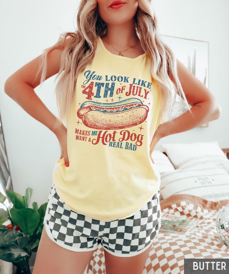 Puede incluir: Camiseta sin mangas amarilla con la frase "You look like the 4th of July makes me want a hot dog real bad." Combinada con shorts a cuadros blancos y negros. El tema general es patri&oacute;tico e informal.