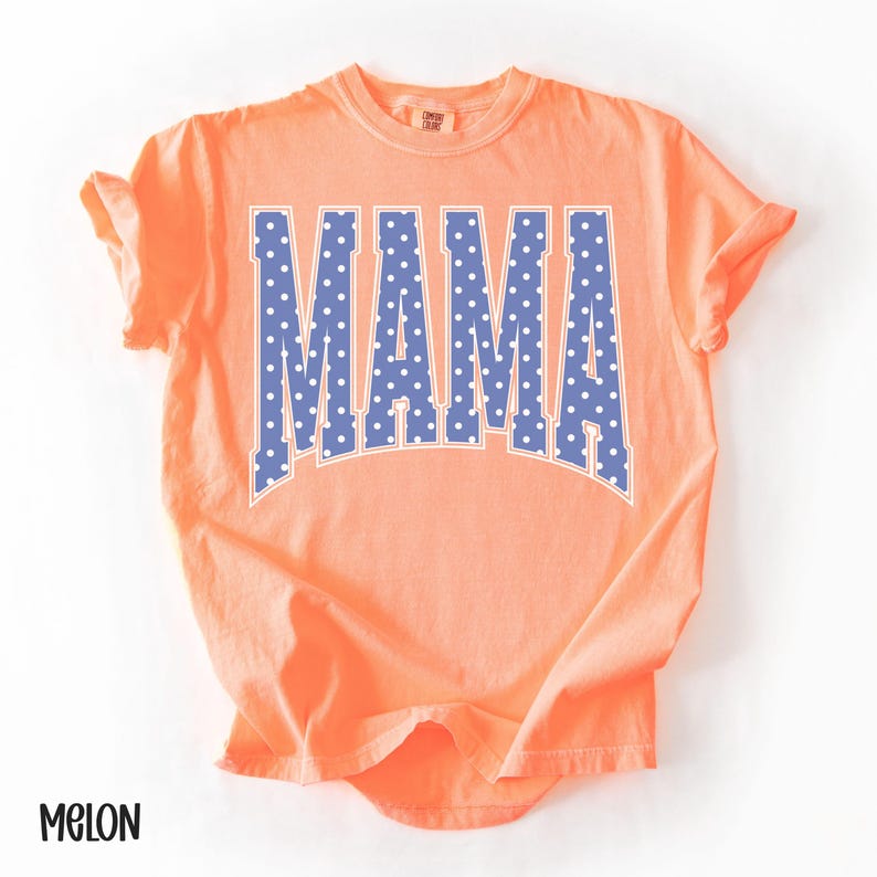 May include: A melon-colored t-shirt with the word "MAMA" in large, blue, polka-dotted letters across the front. The shirt has short sleeves and a crew neck. The word "MELON" is printed in black at the bottom left corner.