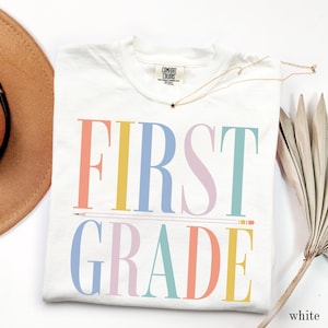 May include: A white t-shirt featuring the text FIRST GRADE in large, multicolored serif letters. The word FIRST is in coral, yellow, blue, lavender, and teal, while GRADE is in blue, lavender, teal, coral, and yellow. A pencil graphic separates the two lines.
