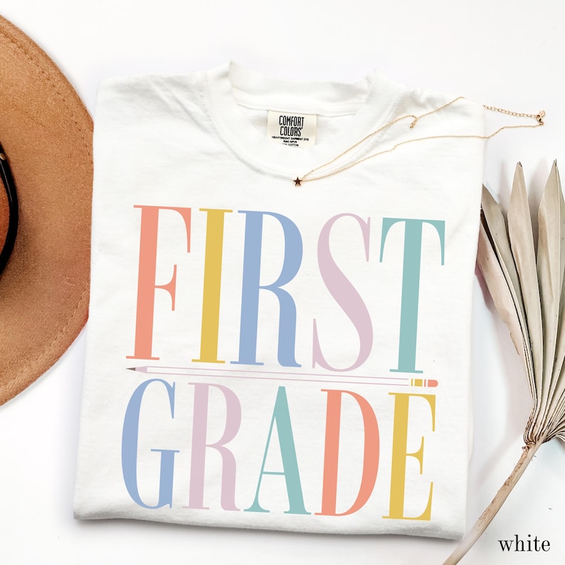 May include: A white t-shirt featuring the text FIRST GRADE in large, multicolored serif letters. The word FIRST is in coral, yellow, blue, lavender, and teal, while GRADE is in blue, lavender, teal, coral, and yellow. A pencil graphic separates the two lines.
