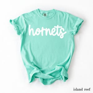 May include: A light turquoise t-shirt with the word "hornets" in white script across the front. The shirt is made of soft material and has rolled-up sleeves. The color is labeled "island reef".