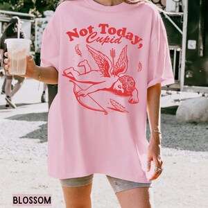 May include: Pink oversized t-shirt with a graphic of a red cupid with wings and an arrow. The text "Not Today, Cupid" is printed in red on the shirt.