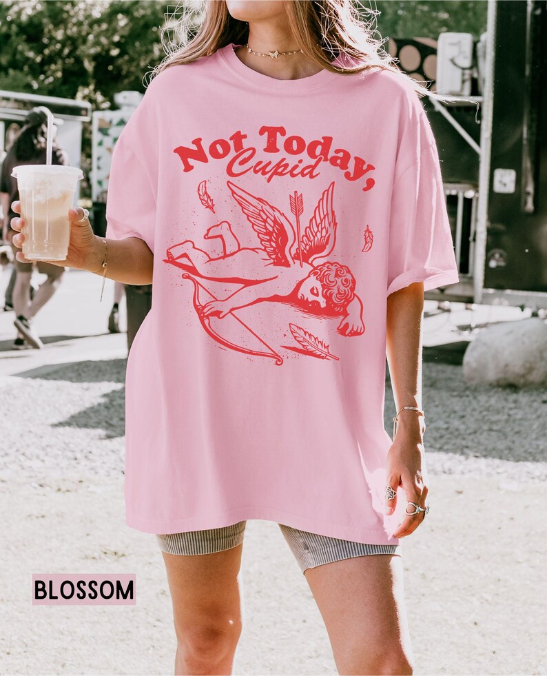 May include: Pink oversized t-shirt with a graphic of a red cupid with wings and an arrow. The text "Not Today, Cupid" is printed in red on the shirt.