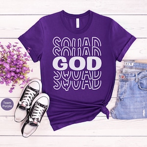 Pu&ograve; includere: Maglietta viola con il testo "GOD SQUAD" in lettere bianche con contorno. Il testo &egrave; ripetuto pi&ugrave; volte in uno schema impilato.