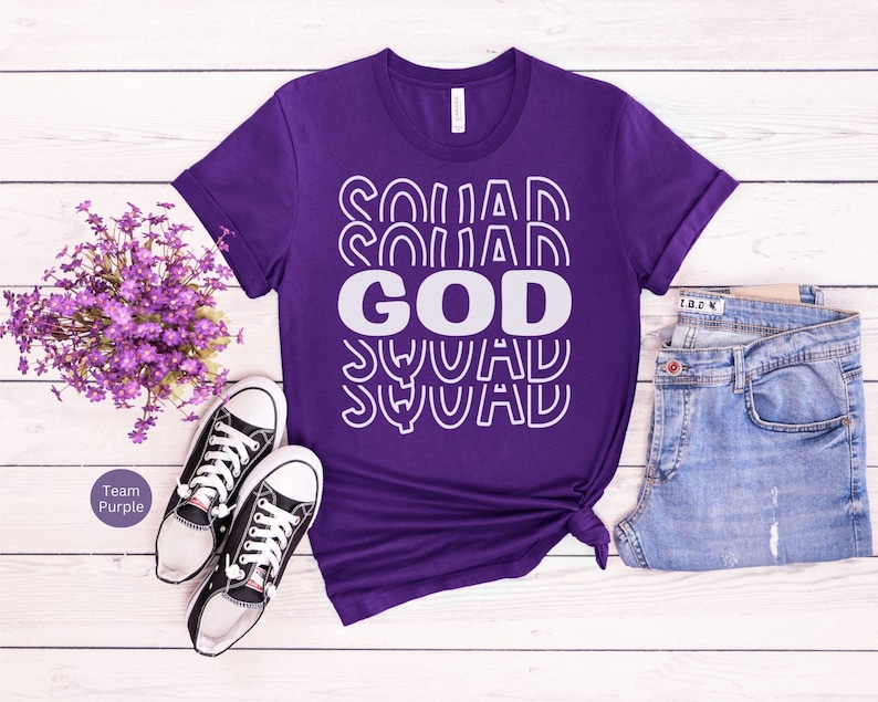 Pu&ograve; includere: Maglietta viola con il testo "GOD SQUAD" in lettere bianche con contorno. Il testo &egrave; ripetuto pi&ugrave; volte in uno schema impilato.
