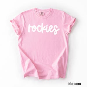 May include: A light pink t-shirt with the word "rockies" in white script across the front. The sleeves are cuffed. The word "blossom" is printed in a small font at the bottom right corner.