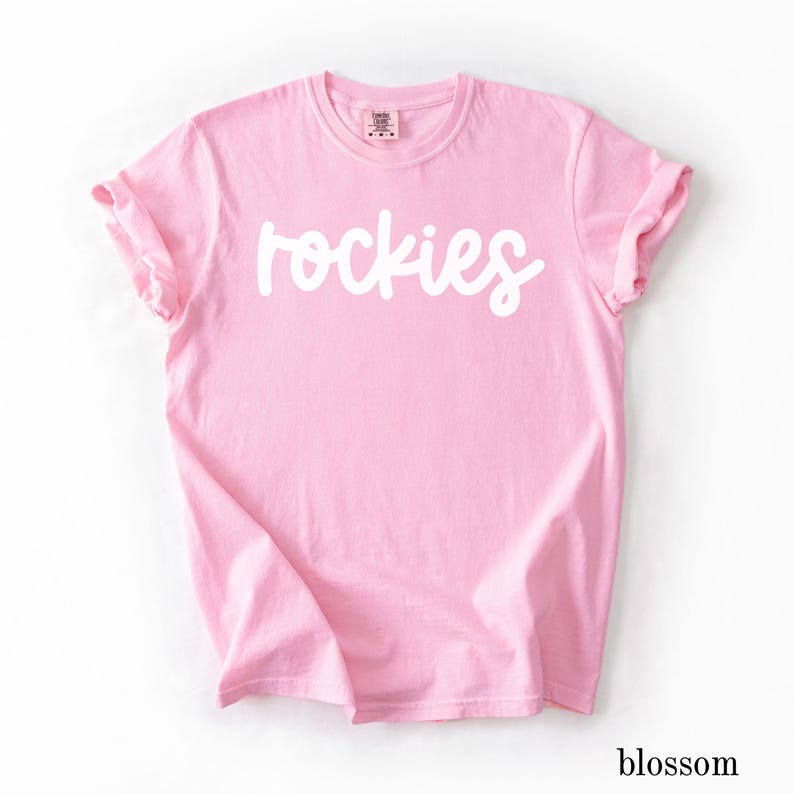 May include: A light pink t-shirt with the word "rockies" in white script across the front. The sleeves are cuffed. The word "blossom" is printed in a small font at the bottom right corner.