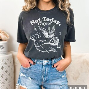 May include: A dark gray t-shirt with a white graphic of a winged cupid with a bow and arrow. The text "Not Today, Cupid" is printed on the shirt.