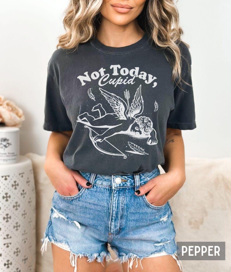 May include: A dark gray t-shirt with a white graphic of a winged cupid with a bow and arrow. The text "Not Today, Cupid" is printed on the shirt.