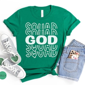 Pu&ograve; includere: Una maglietta verde con il testo "GOD SQUAD" in lettere bianche con un design retr&ograve;. La maglietta &egrave; stesa su uno sfondo bianco con un paio di scarpe da ginnastica grigie e pantaloncini di jeans blu.